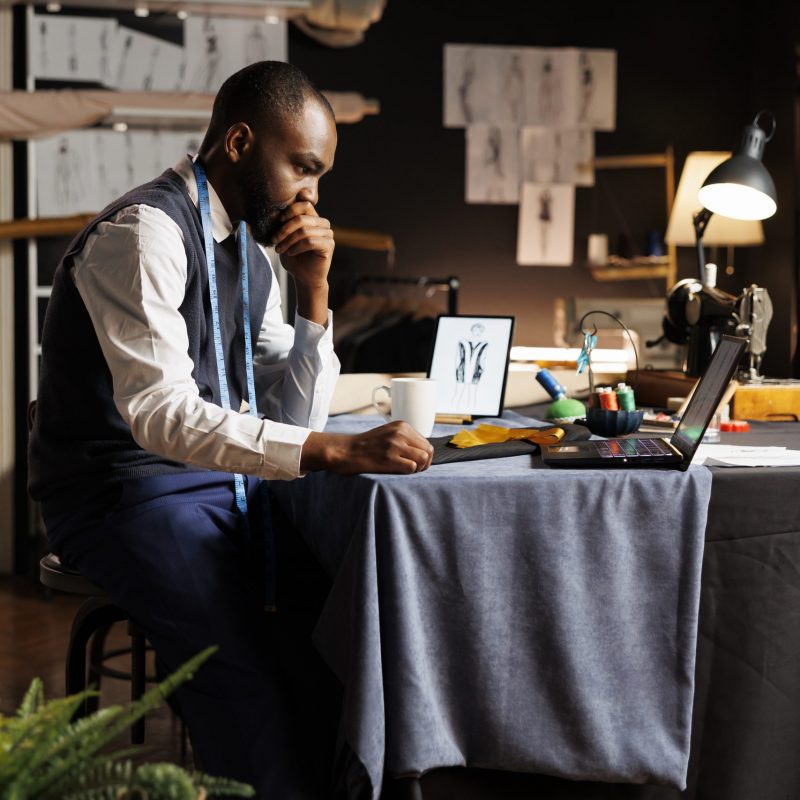 Couturer looking online for inspiration, struggling to find ideas for upcoming fashion design runway show collection. Suitmaker working on bespoken sartorial garments in atelier shop workspace